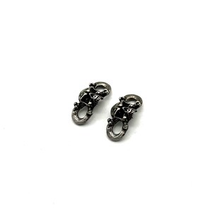 May include: Two small, silver-toned skull-shaped clasps. Each clasp features a detailed skull design with an open mouth and eye sockets. The clasps have a loop at the bottom for attachment. The background is plain white.