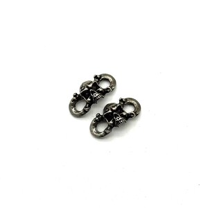 May include: Two silver-toned skull-shaped connectors. Each connector features a detailed skull design with open eye sockets and teeth. The connectors are linked by a circular loop, suggesting they are designed to connect two items. The metal has a slightly aged appearance.