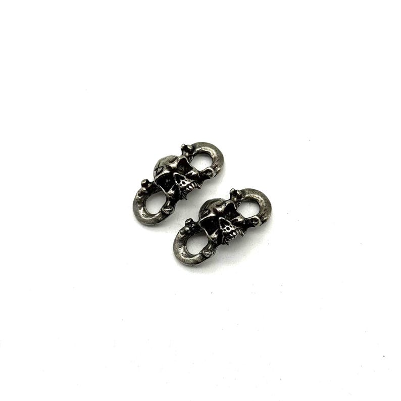 May include: Two silver-toned skull-shaped connectors. Each connector features a detailed skull design with open eye sockets and teeth. The connectors are linked by a circular loop, suggesting they are designed to connect two items. The metal has a slightly aged appearance.