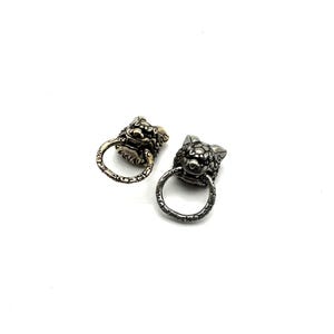 May include: Two metal lion head charms with ring handles. One charm is gold-toned, the other is silver-toned. The lion heads have detailed features and are set against a white background. These charms could be used for jewelry making or other crafts.
