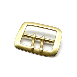 May include: A gold-toned metal buckle with a rectangular shape and two prongs. The buckle is designed for use with a belt or strap.