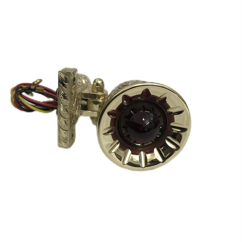 May include: A gold motorcycle taillight with a red lens and a decorative design. The taillight has a mounting bracket and wires.