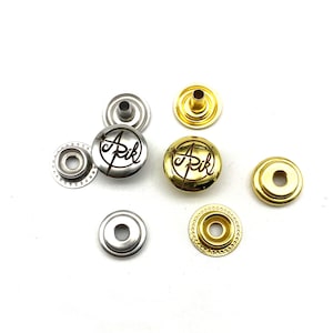 Puede incluir: Un conjunto de botones a presión de metal dorado y plateado con un logotipo que dice "A Pik" en una fuente cursiva. Los botones se utilizan para la ropa y otras manualidades.