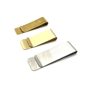 Op de afbeelding: Drie metalen geldclips, een gouden, een messingkleurige en een zilveren. De clips zijn allemaal even groot en van dezelfde vorm, en ze zijn in een rij van links naar rechts gerangschikt.