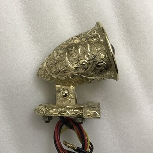 May include: A gold-colored, ornate, decorative light with a floral design. The light has a round, black lens and a base with three mounting holes.