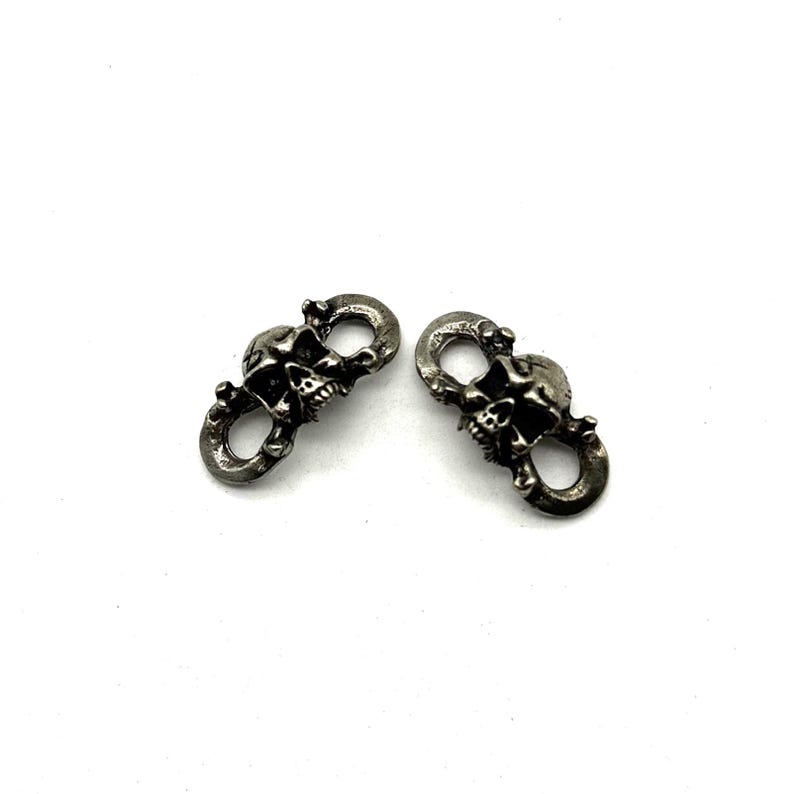 May include: Two silver-colored skull-shaped clasps. Each clasp features a detailed skull design with open eye sockets and teeth. The clasps have a loop at the top and bottom for attachment. The background is plain white.