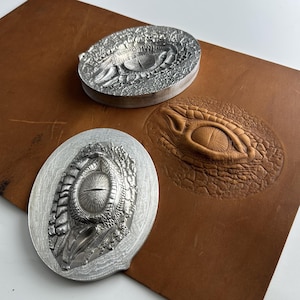Puede incluir: Dos sellos metálicos con diseños detallados de ojos de reptil, uno ovalado y otro redondo, junto a una pieza de cuero con un ojo en relieve. Los sellos son plateados y el ojo en relieve es marrón.