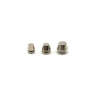 Può includere: Tre piccoli oggetti metallici color argento di forma conica. Ogni oggetto ha una parte superiore piatta e una base leggermente più larga. Gli oggetti sono disposti in fila su uno sfondo bianco.