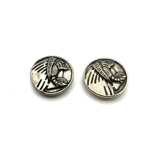 Puede incluir: Dos botones redondos de metal con un diseño de tocado nativo americano. Un botón es plateado y el otro es dorado.