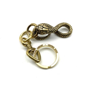 May include: A gold-toned brass key chain with a snake design. The snake is coiled into an infinity symbol. The key chain has a split ring and a swivel clasp. The key chain is engraved with "Leather House Fieldwork Company".