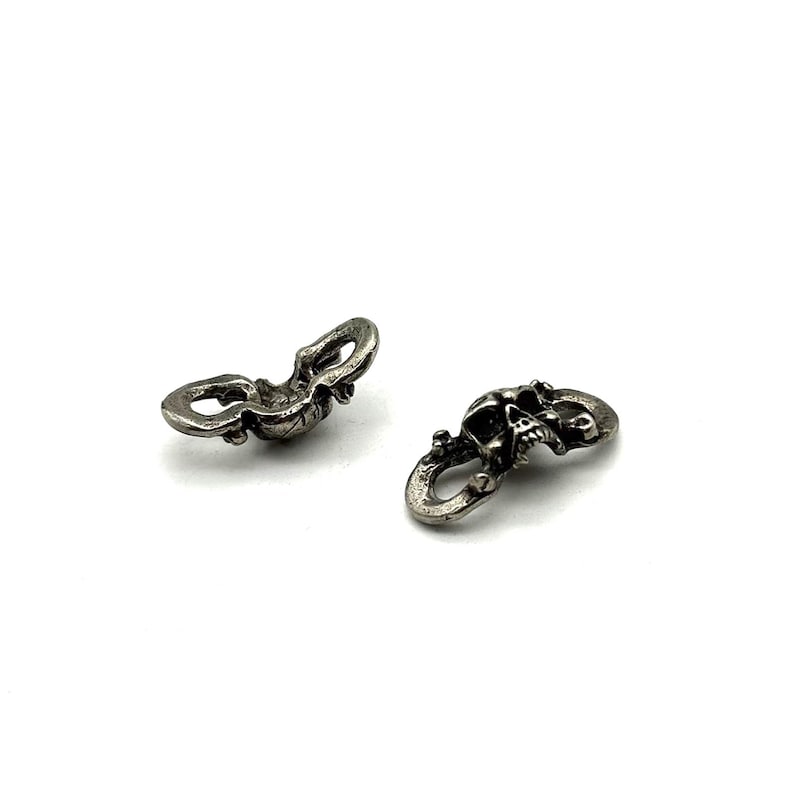 May include: Two small, silver-colored skull charms. Each charm features a detailed skull design with a loop at each end. The charms are displayed on a white background, showcasing their intricate details and metallic finish.