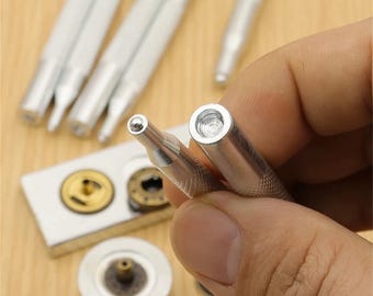 May include: Close-up of several silver metal tools, including a punch and a setting tool, used for installing rivets or snaps. The tools have textured handles for grip and are designed for precision work. Other metal components are also visible.