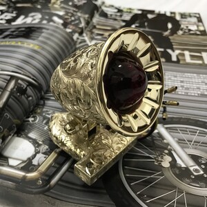May include: A gold-colored motorcycle taillight with an ornate, engraved design. The taillight has a red glass lens and a decorative, fluted rim.
