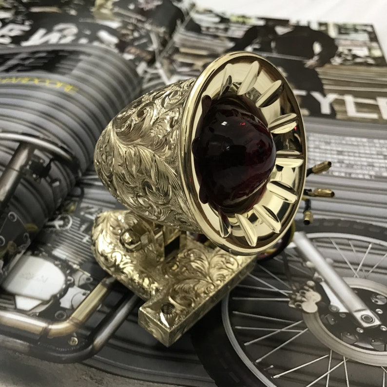 May include: A gold-colored motorcycle taillight with an ornate, engraved design. The taillight has a red glass lens and a decorative, fluted rim.