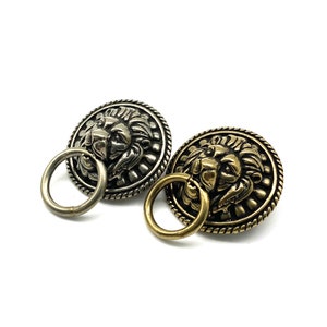 May include: Two ornate lion head buttons with a ring attached. One button is silver and the other is gold.