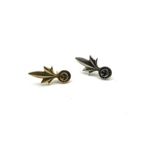 May include: Two small metal pins in the shape of a stylised flame with a round disc at the end. One pin is gold and the other is silver.