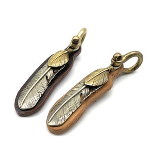 以下が含まれることがあります： 羽のデザインが施された2つの革製キーホルダー。1つは濃い茶色で、銀色の羽と金色の羽のアクセントが付いています。もう1つは明るい茶色で、銀色の羽と金色の羽のアクセントが付いています。どちらも真鍮色のループが付いています。