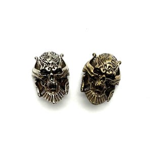 Op de afbeelding: Twee schedelvormige ringen, een zilveren en een gouden, met ingewikkelde details en een gotisch ontwerp.