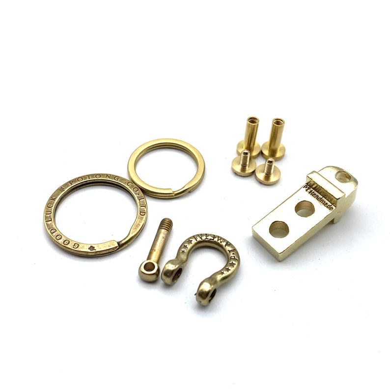 May include: A set of brass key chain components including a key ring, a small hook, a rectangular piece with two holes, and three screws. The key ring has the text "GOOD LUCK POITONG CO LTD" engraved on it. The hook has the text "NEW WAY" engraved on it. The rectangular piece has the text "Handmade" engraved on it.