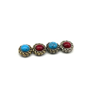 Pode incluir: Quatro botões decorativos redondos com uma pedra central em turquesa e vermelho. As pedras são fixadas numa moldura de metal com uma borda em forma de corda. Os botões estão sobre um fundo branco.