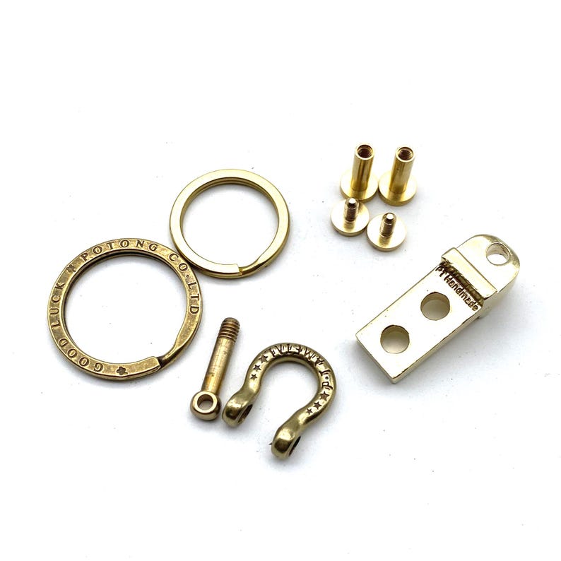 May include: A set of brass key chain hardware, including a key ring, a D-ring, a hook, and two screws. The key ring has the text "LUCKY POTONG CO. LTD GOOD" engraved on it. The hook has the text "FULL METAL JACKET" engraved on it. The hardware is handmade.