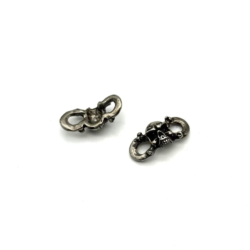 May include: Two small, silver-colored skull charms. Each charm features a skull design with intricate details and is connected to two looped ends. The charms are set against a white background.