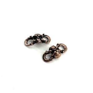 Puede incluir: Dos cierres en forma de calavera de color bronce. Cada cierre presenta un diseño detallado de calavera con bucles curvos a cada lado. Los cierres están sobre un fondo blanco, resaltando sus intrincados detalles y acabado metálico.
