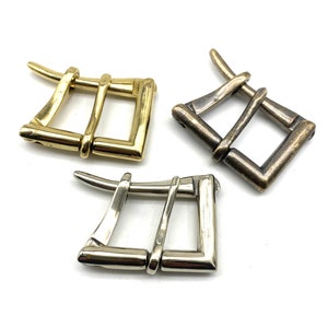 May include: Three metal belt buckles in different finishes: gold, silver, and antique brass. Each buckle has a unique, intricate design with a square shape and two loops.