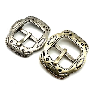 Pode incluir: Duas fivelas decorativas de metal. Uma é prateada, a outra dourada. Ambas têm um design ornamentado e gravado com formato quadrado e uma barra central para a correia do cinto. As fivelas estão sobre um fundo branco.