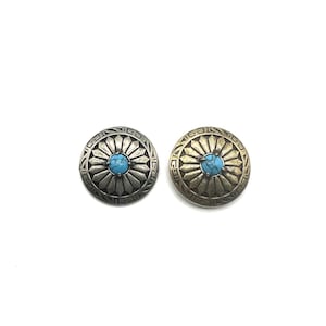 May include: Two round metal buttons with a turquoise stone in the centre. The buttons have a decorative design around the stone, with one button in silver and the other in gold.