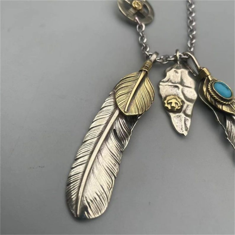 May include: A silver chain necklace with multiple pendants. The pendants include a silver feather with a gold feather overlay, a silver and gold skull, and a silver feather with a turquoise stone.