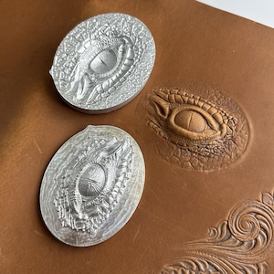 Puede incluir: Dos sellos de metal plateado con un diseño detallado de ojo de cocodrilo, junto a una pieza de cuero marrón con un ojo de cocodrilo en relieve. Los sellos son ovalados y muestran escamas y detalles de ojo intrincados. El cuero tiene un ojo en relieve similar.