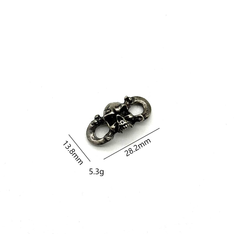 May include: A silver-colored metal clasp featuring a skull design. The clasp measures 28.2mm in length and 13.8mm in width, with a weight of 5.3 grams. The skull is centered between two circular loops.