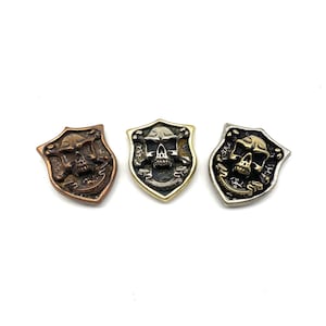 Op de afbeelding: Drie metalen schildspelden met een schedel en gekruiste beenderen in verschillende afwerkingen: koper, messing en zilver.