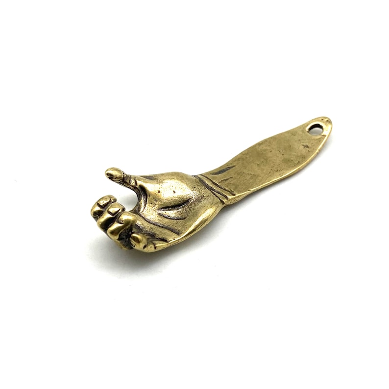 May include: A brass bottle opener shaped like a hand with a thumb pointing up. The opener has a flat, rectangular blade.