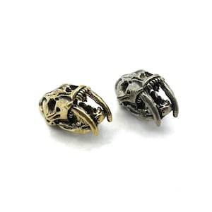 Pode incluir: Dois pingentes de metal em forma de crânio de tigre dente de sabre, um em ouro e outro em prata. Os pingentes são detalhados e realistas, com dentes afiados e uma expressão feroz.
