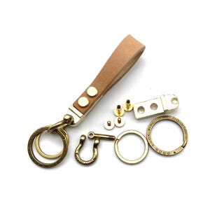 May include: A brass key chain kit with a tan leather strap, two brass rings, a brass hook, and a brass plate with the text "GOOD LUCK" and "SPOTONG CO. LTD".