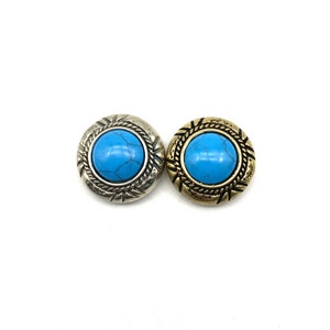 Op de afbeelding: Twee ronde turquoise cabochon-knoppen met sierlijke zilveren en gouden metalen omlijstingen. De knoppen hebben een decoratief touwontwerp rond de turquoise stenen.