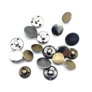 May include: A collection of metal snap fasteners in various finishes, including silver, bronze, and black. The snaps are in different sizes and shapes, with both the button and socket components visible. The fasteners are scattered on a white background.