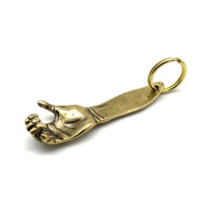 May include: A brass bottle opener shaped like a hand with a ring attached. The hand is open and the fingers are curled to hold a bottle cap.
