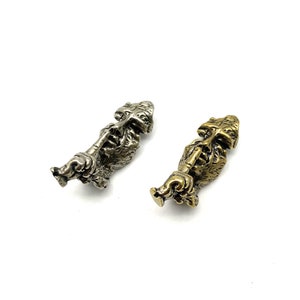 Op de afbeelding: Twee kleine metalen sculpturen van een gezicht met een vinger over de mond, een is zilver en de andere is goud.