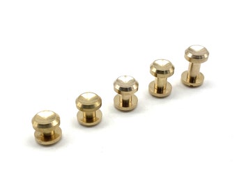 May include: Five brass thumb tacks of varying sizes, arranged in a row from smallest to largest on a white surface.