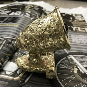 May include: A gold-colored motorcycle headlight with an intricate floral design. The headlight is mounted on a base with a similar design.