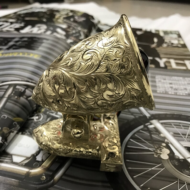May include: A gold-colored motorcycle headlight with an intricate floral design. The headlight is mounted on a base with a similar design.