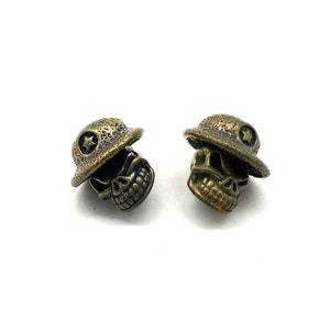 May include: Two bronze-colored skull beads, each wearing a hat with a star emblem. The skulls have detailed features, including teeth and eye sockets. The hats are slightly tilted, adding a playful touch to the design. The background is plain white.