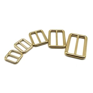 May include: Five gold-toned metal belt buckle slides of varying sizes. The buckles are rectangular with a center bar.