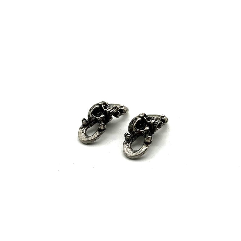 May include: Two small, silver-toned skull-shaped charms. Each charm features a detailed skull design with an open loop at the bottom. The charms are displayed against a white background.