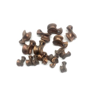 May include: A collection of small, bronze-coloured metal charms in the shape of a stylised knot. The charms are arranged in a random pattern on a white background.