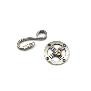 May include: Two-piece silver-toned jewelry findings set. One piece is a hook with a snake head design. The other is a circular piece with a gold-toned center and square accents. The set is on a white background.