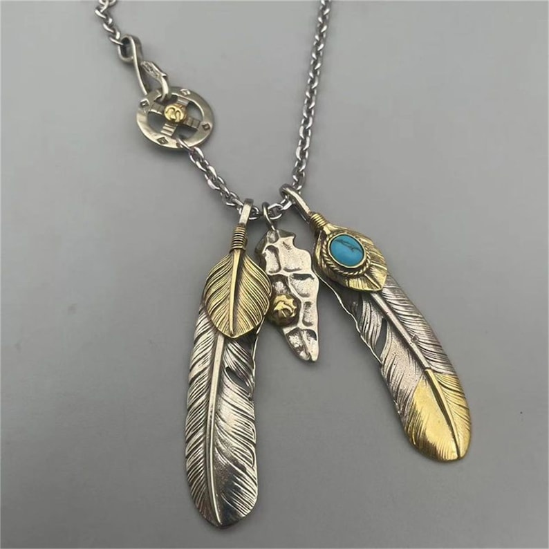 May include: A silver chain necklace with a silver and gold feather pendant. The pendant features three feathers, one with a turquoise stone.